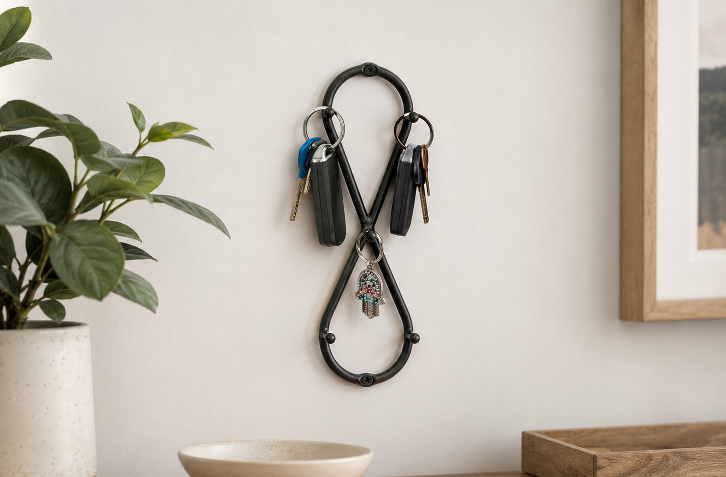 The Vertical Infinity wrought iron key holder mounted on a light-colored wall with several sets of keys hanging from the infinity-symbol frame, flanked by a textured bowl and vase
