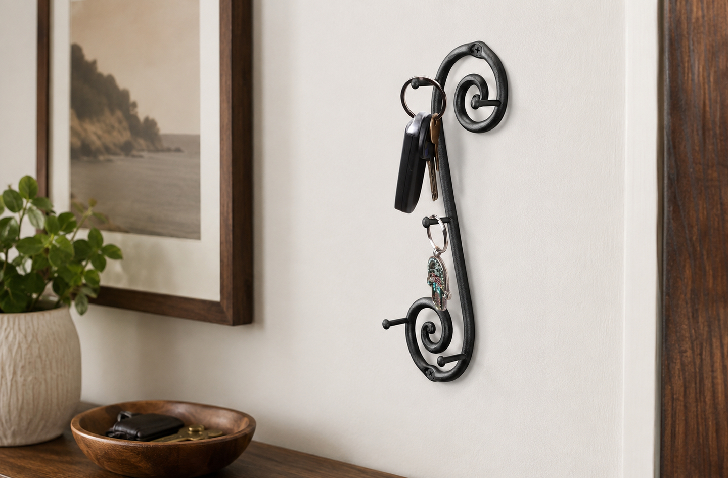 RTZEN wrought iron vertical wall-mounted key holder holding two sets of keys against an off-white wall beside a framed coastal print