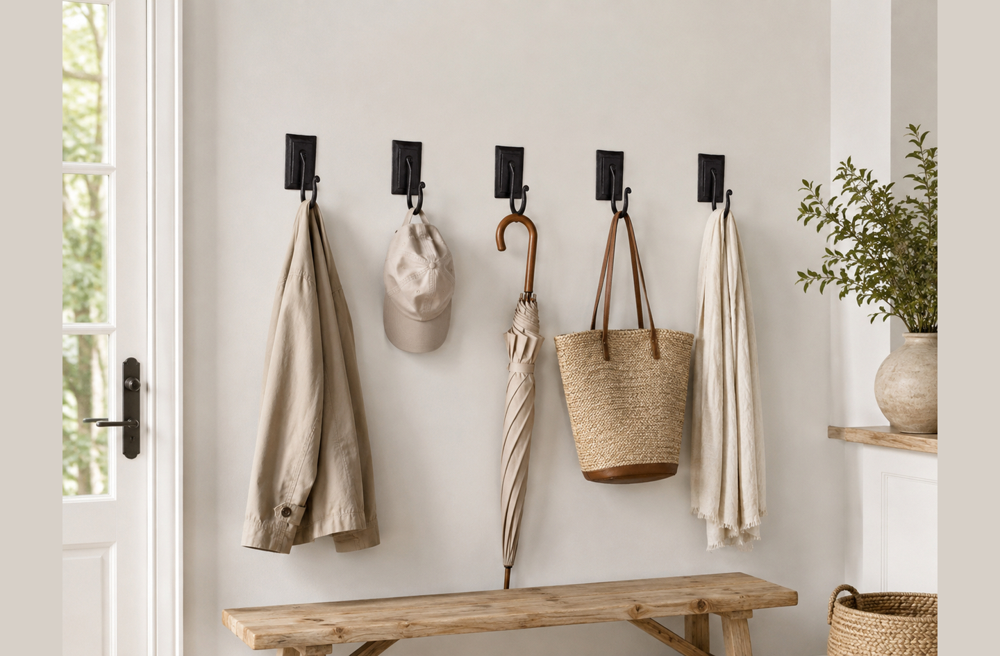 Five wrought iron double plate wall hooks mounted in a row above a wooden bench, each holding a different everyday item including a coat, a cap, an umbrella, a tote bag, and a scarf