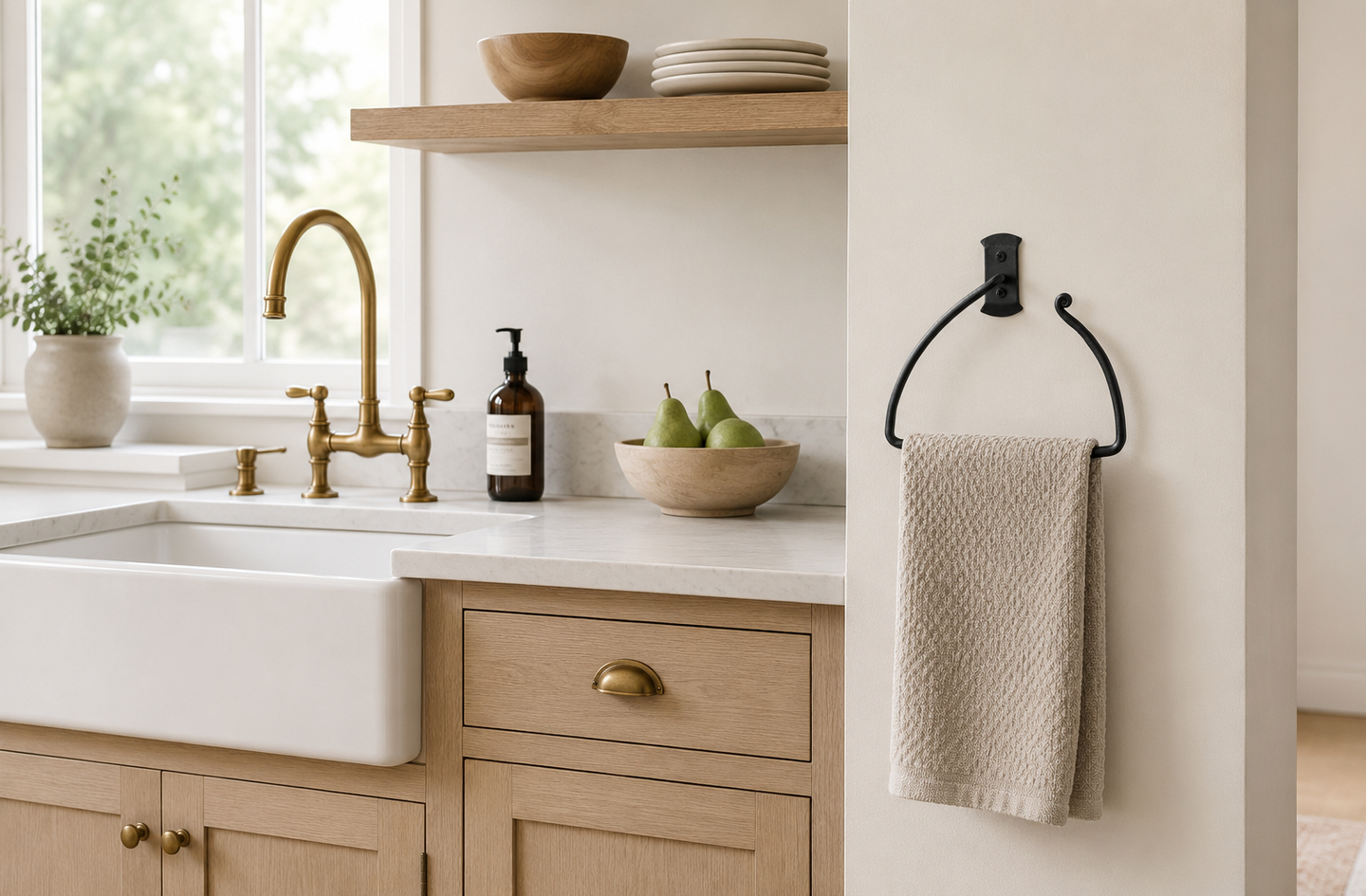 A black half-circle wrought iron hand towel holder with a light beige towel mounted on a white wall next to a farmhouse kitchen sink and wooden cabinets.