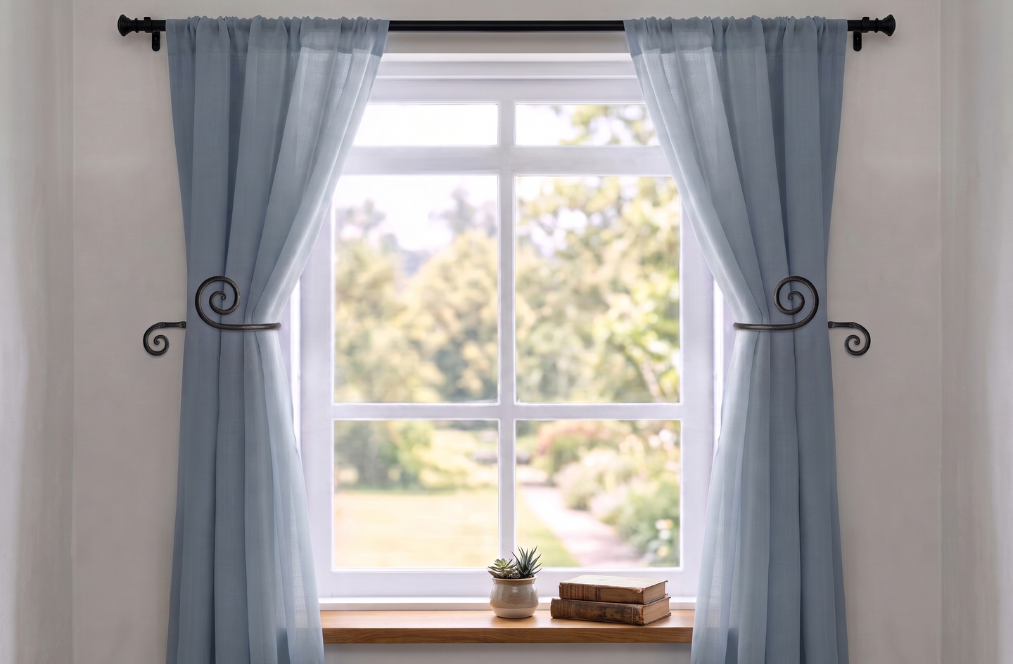 A pair of RTZEN black wrought iron swirly curtain holdbacks securing light blue curtains against a window frame, beside a windowsill with a small succulent and two stacked vintage books.