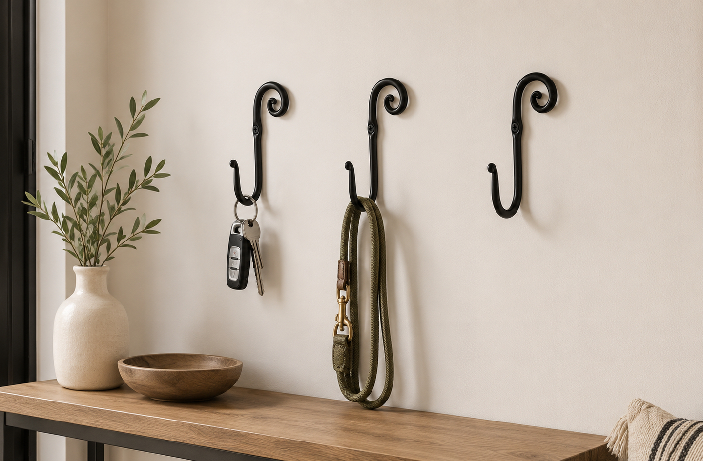 Three decorative black wrought iron wall hooks mounted on a plain wall above a wooden entryway table, with keys and a dog leash hanging from two of the hooks.