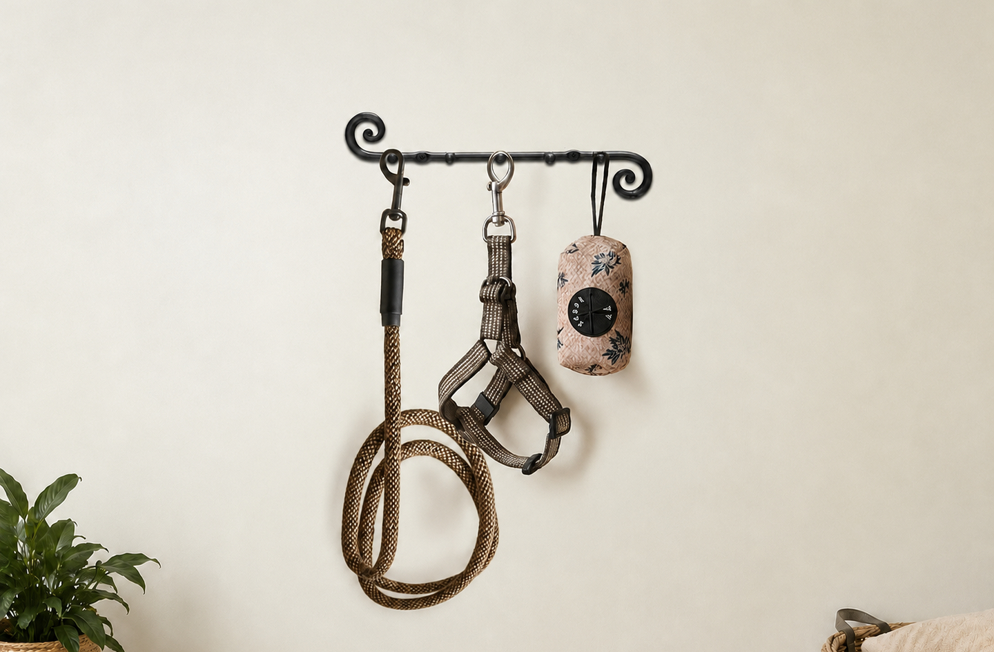 A black wrought iron hook rack mounted on a plain light wall holds a rope dog leash, a harness and a patterned waste bag dispenser, with a small potted plant in the lower corner.