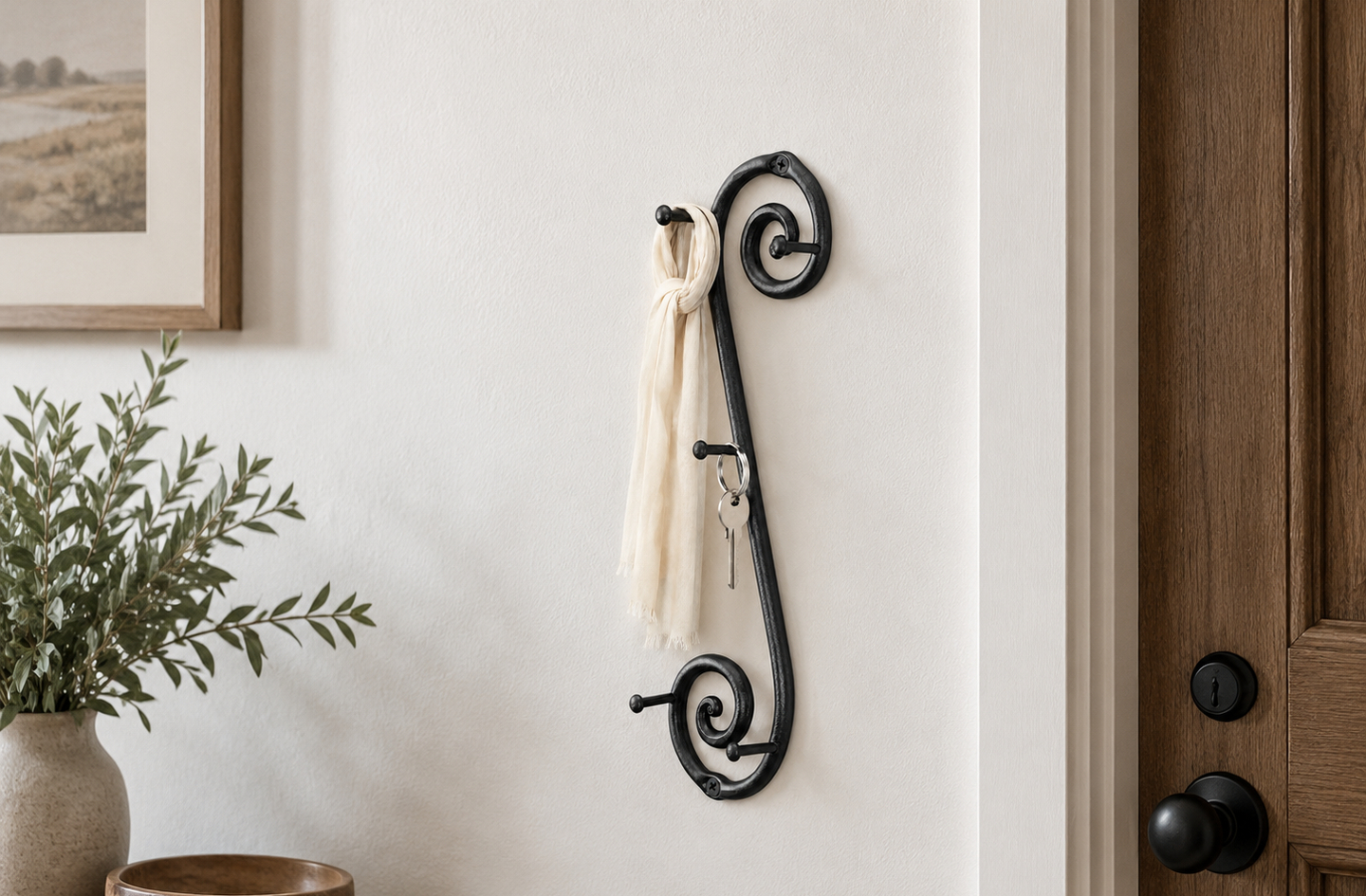 Compact key holder idea for a narrow wall: a black wrought iron vertical key and coat rack mounted beside a wooden entry door, with a framed picture and a vase of greenery to the left.