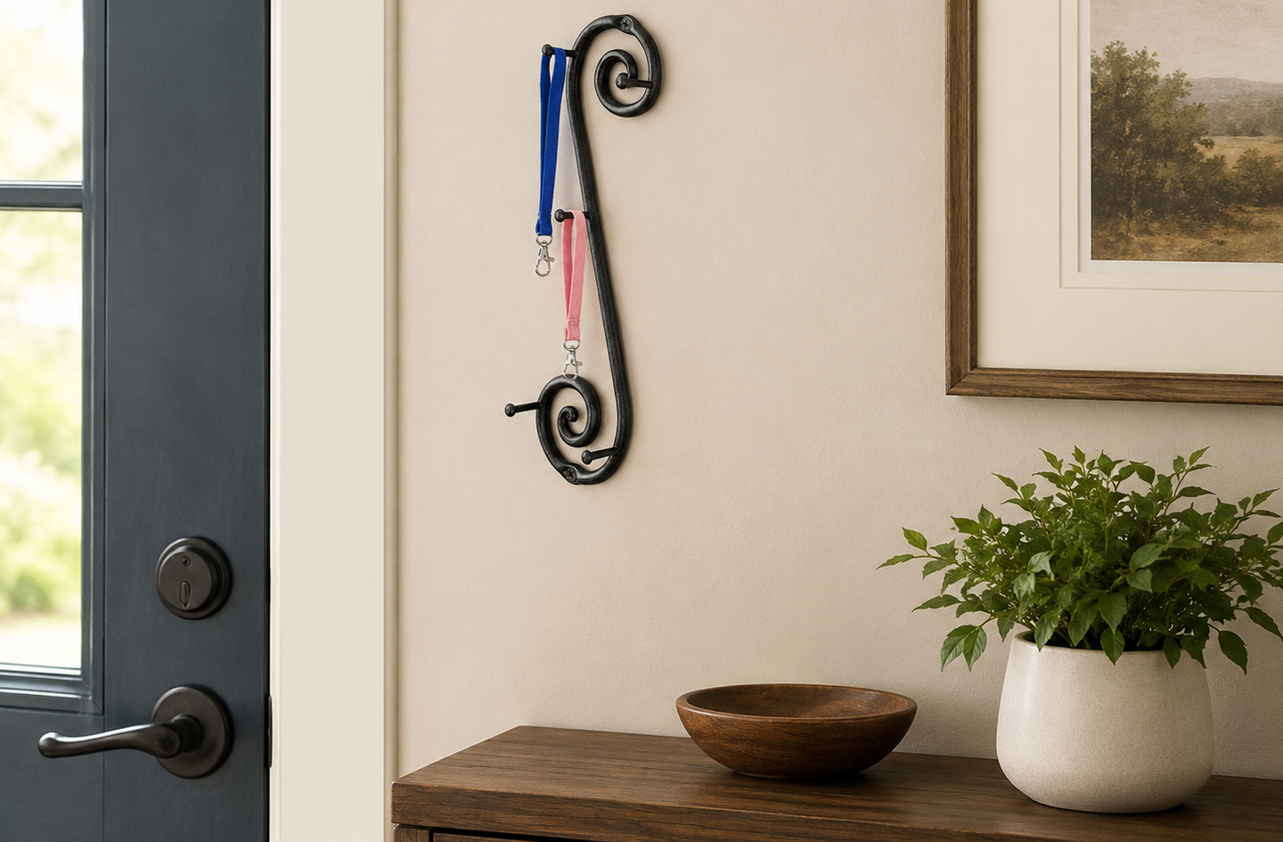 A black wrought iron vertical key hanger on a beige wall holding a blue lanyard and a pink lanyard above a wooden surface with a small bowl and a plant.